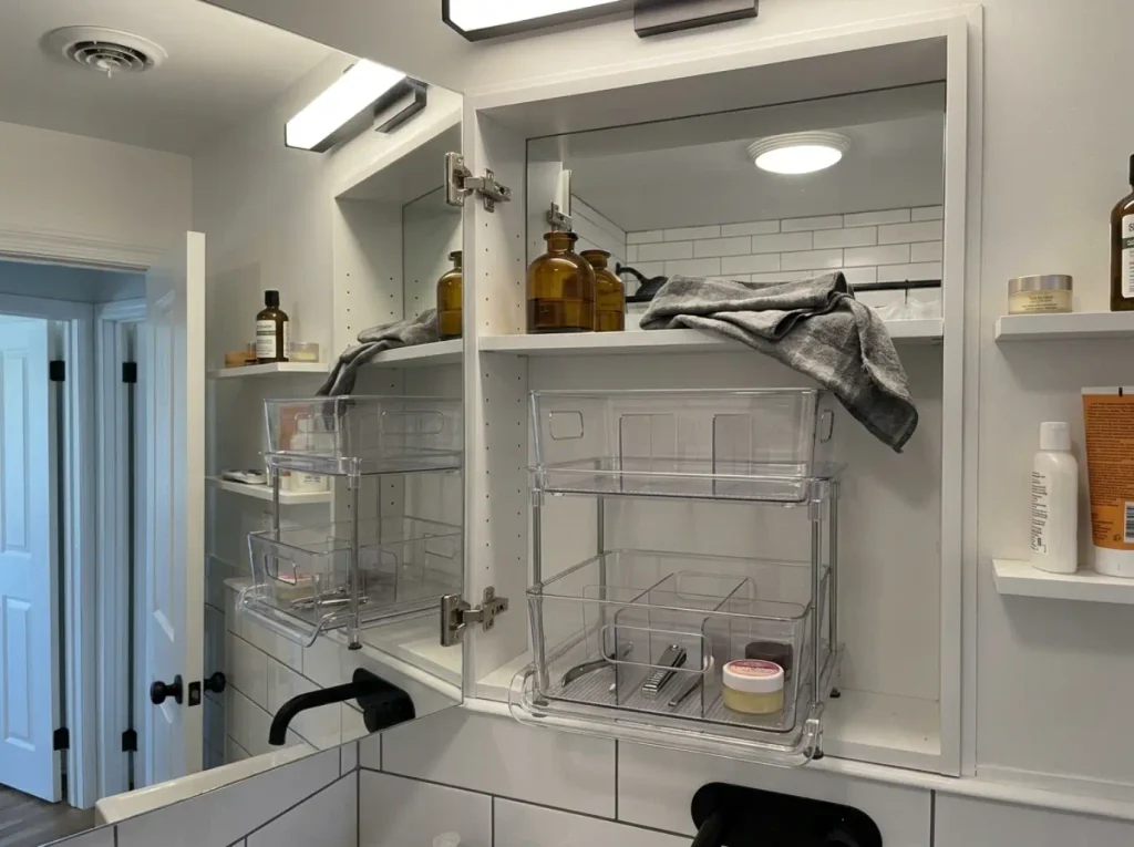 Divided clear acrylic organizer holding tweezers and nail clippers separately on a medicine cabinet shelf