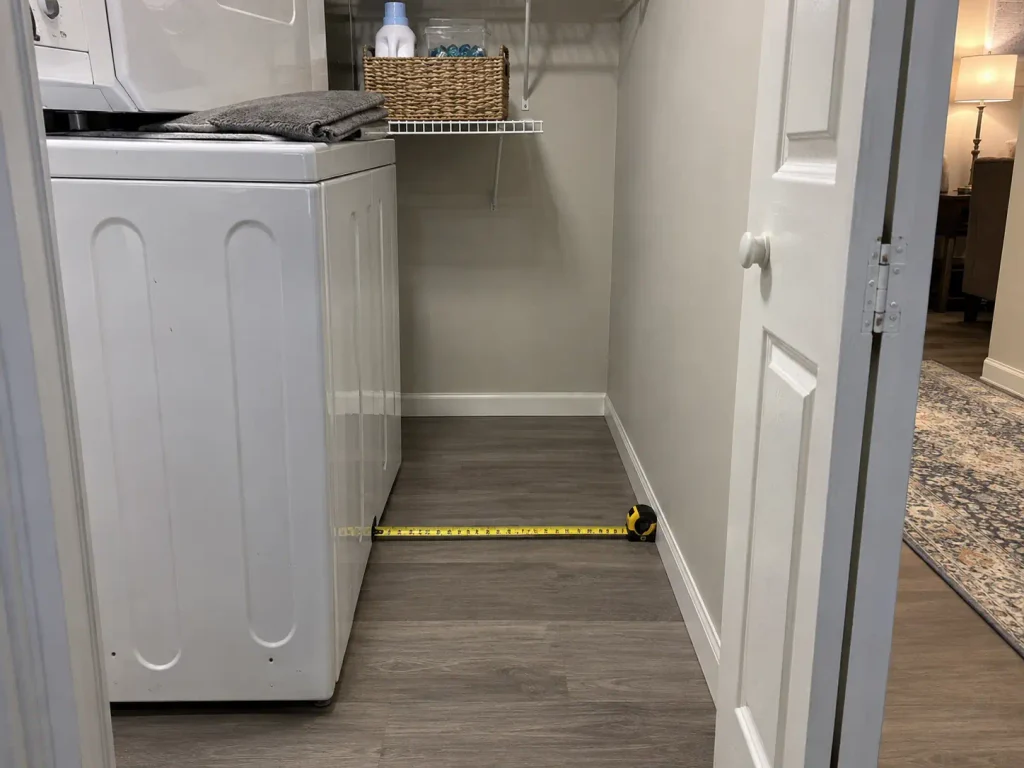 Tape measure extended across the narrow floor gap beside a stacked washer dryer in a small laundry closet