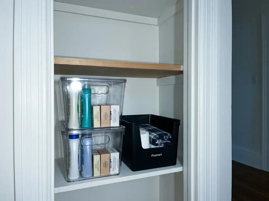 Clear stackable bins holding toiletry backstock on a linen closet shelf, contents visible through the sides