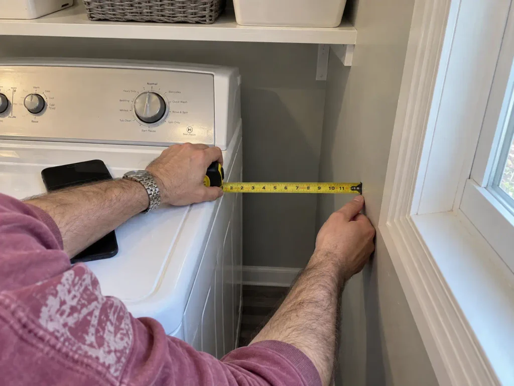 Hands measuring the narrow gap beside a washer with a tape measure before buying laundry room storage