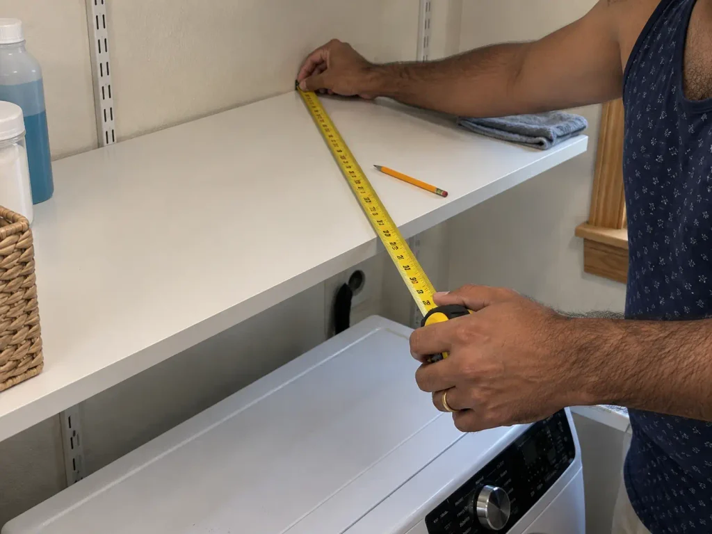 Hands measuring shelf depth above a washer with a tape measure in a small laundry room