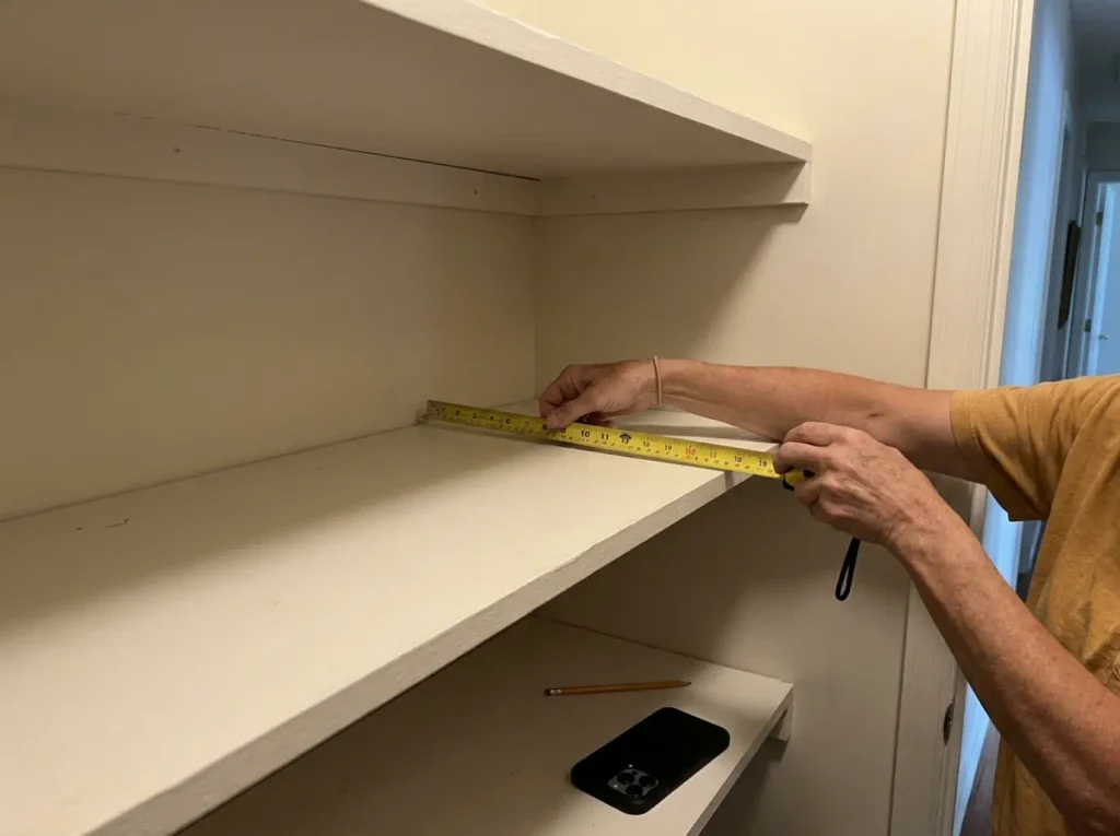 Hands measuring the depth of an empty linen closet shelf with a yellow tape measure