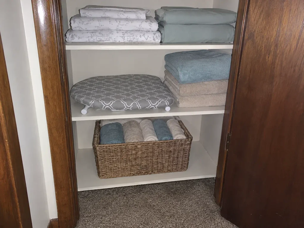 Compact tabletop ironing board folded flat on the shelf of a narrow hallway closet next to towels