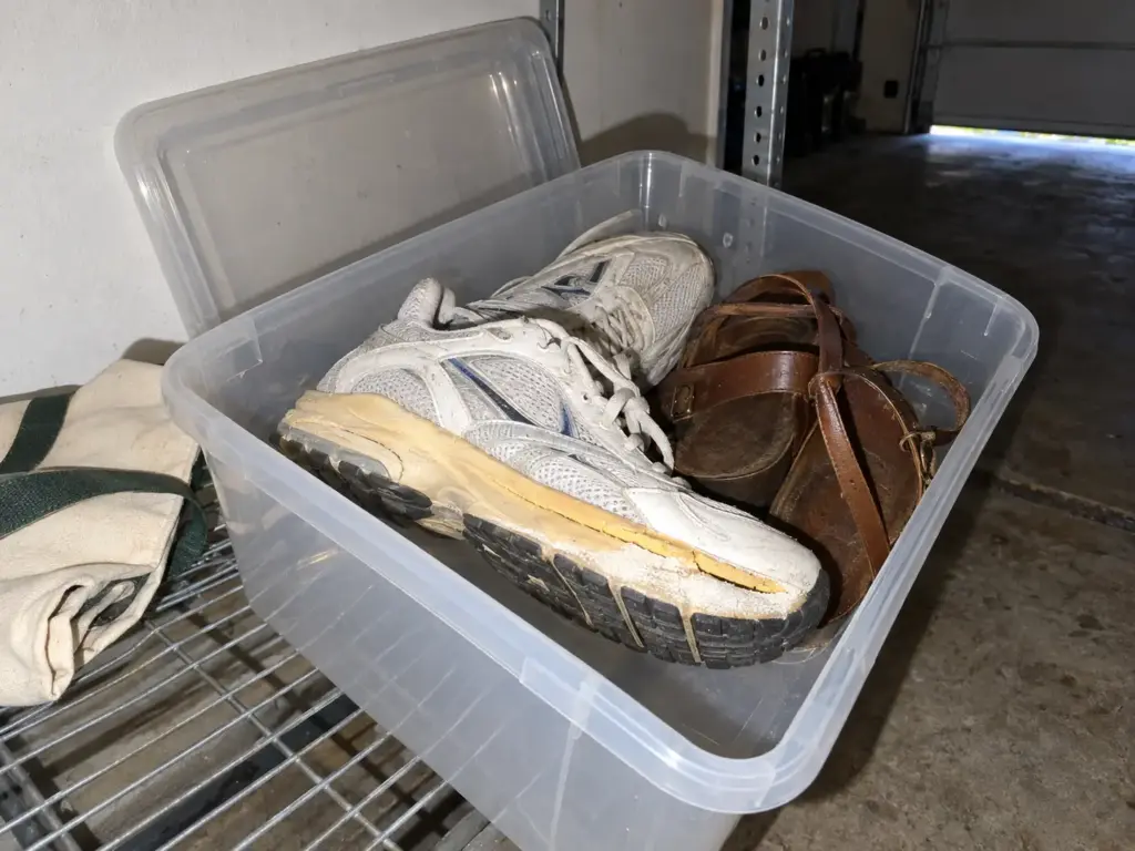 An open tote on a garage shelf holds white sneakers whose midsole edge has yellowed and begun to lift at the toe