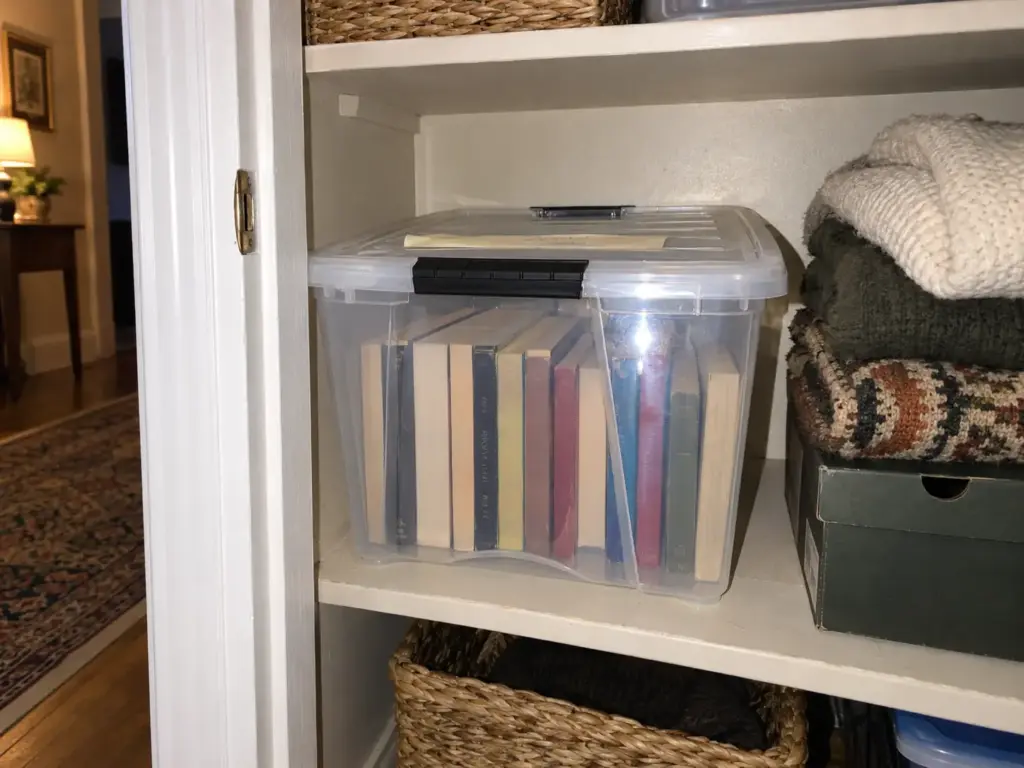A clear IRIS storage box with a lid holding books on a hallway closet shelf, a strip of masking tape across the lid