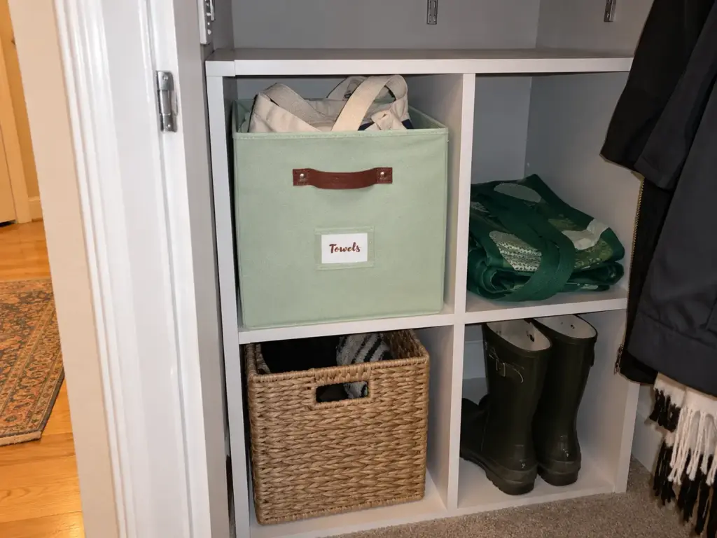 A sage green collapsible fabric bin with a label holder sits on a cube shelf in an open entry closet, a folded tote inside