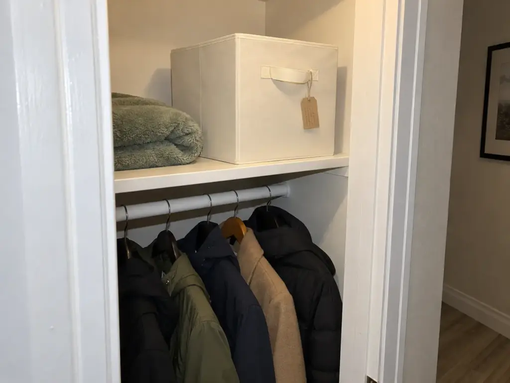 A closed fabric storage cube with a paper tag on its handle sits on a hall closet shelf holding maybe toys