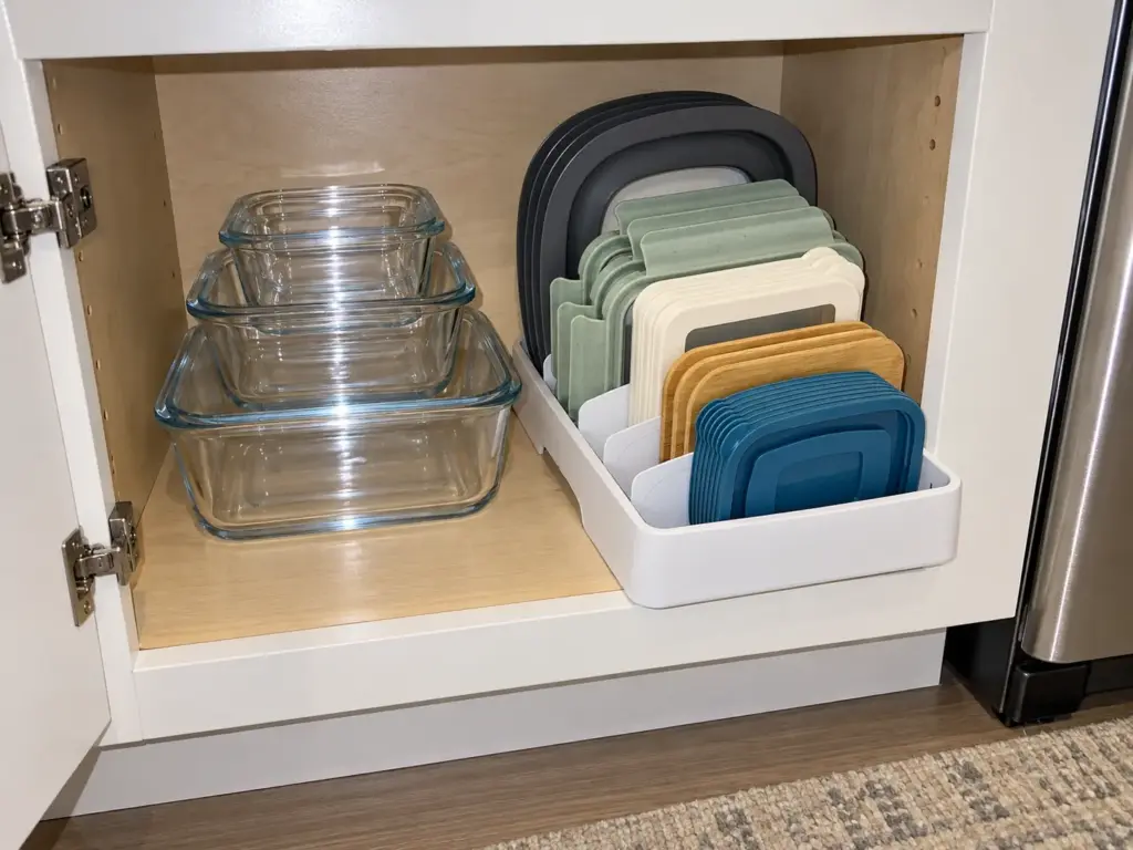 A kitchen cabinet with a lid organizer holding matched lids standing upright beside stacked food containers