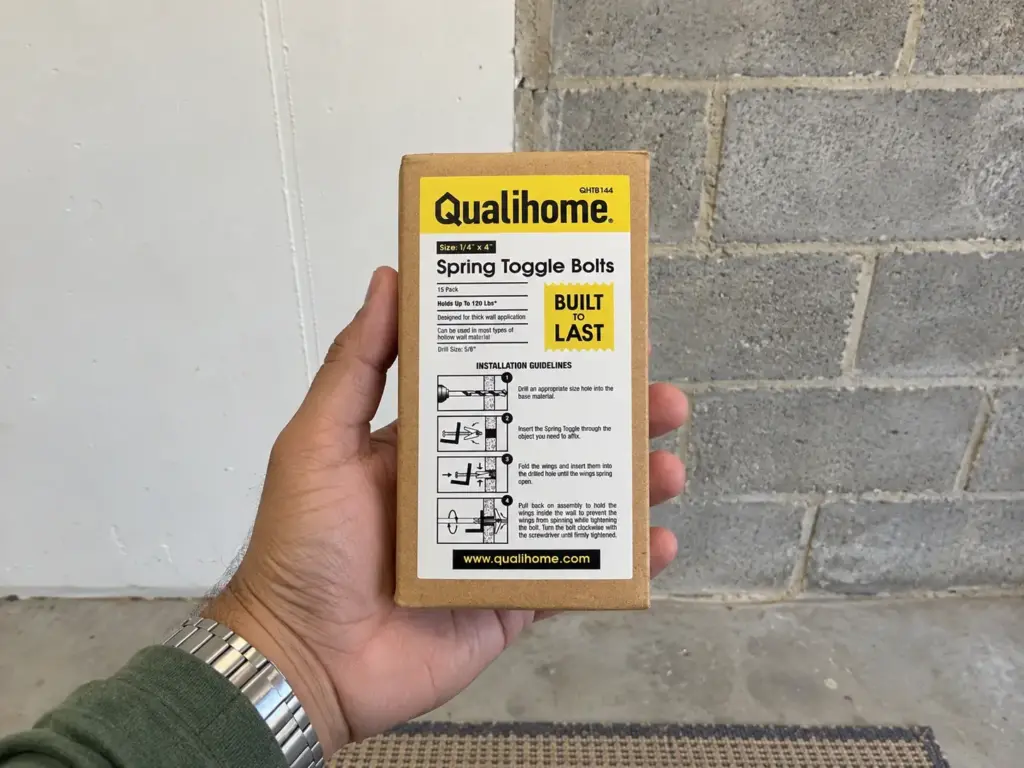Best Garage Storage Systems by What You Actually Store 7 Open palm holding a sealed pack of hollow-wall fasteners in front of a garage corner of white drywall and bare concrete block