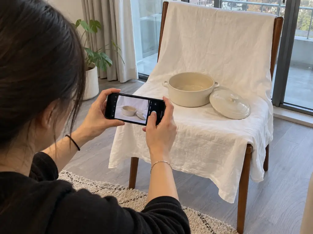 A woman's hands photograph a ceramic casserole dish on a chair draped with a white sheet beside the balcony door