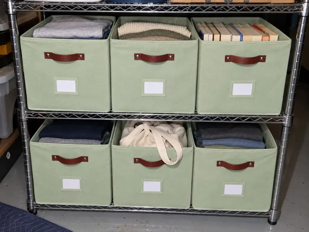 Six sage-green collapsible fabric bins on a garage shelf, each with a blank label card, one bin per donation destination