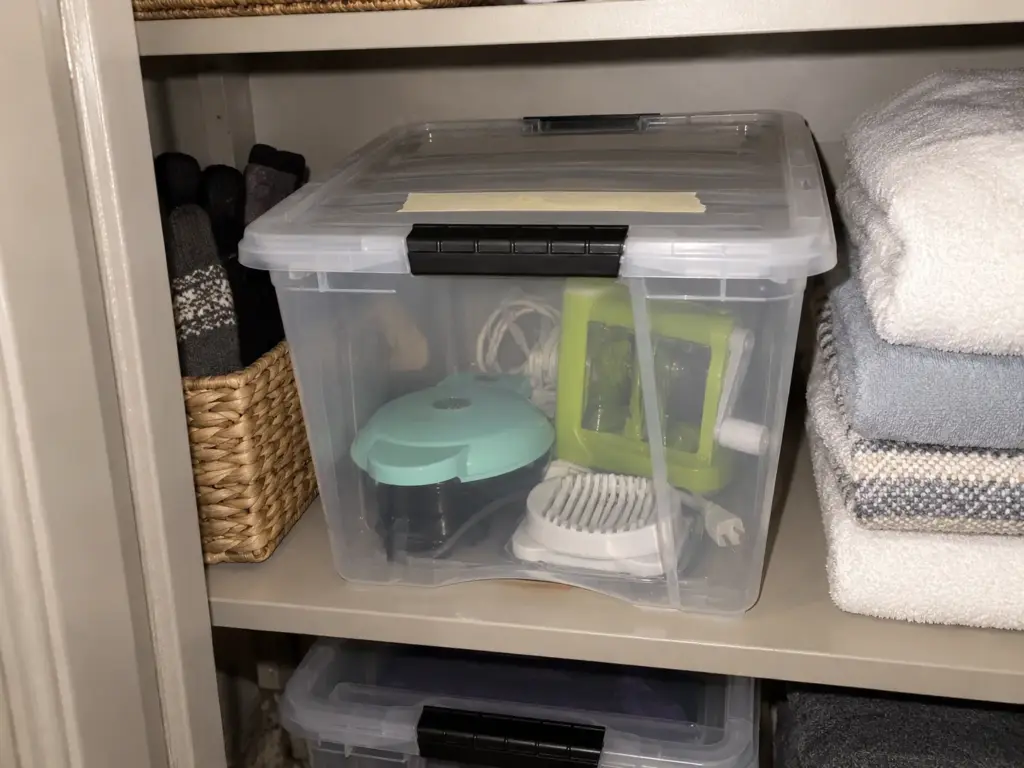 A clear latching box on a hall closet shelf holds a mini waffle maker, an egg slicer and a spiralizer, with tape on the lid