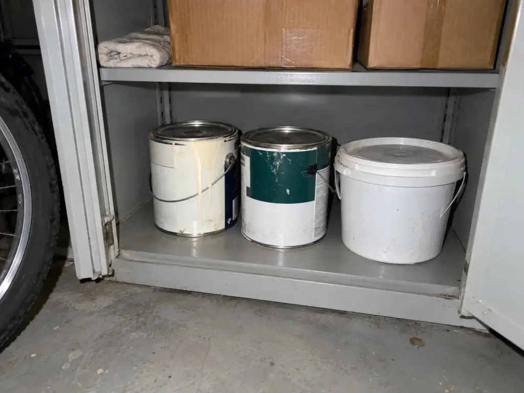 Two closed paint cans and a lidded bucket stand upright on a low garage shelf, lit by flash, waiting for a city drop-off