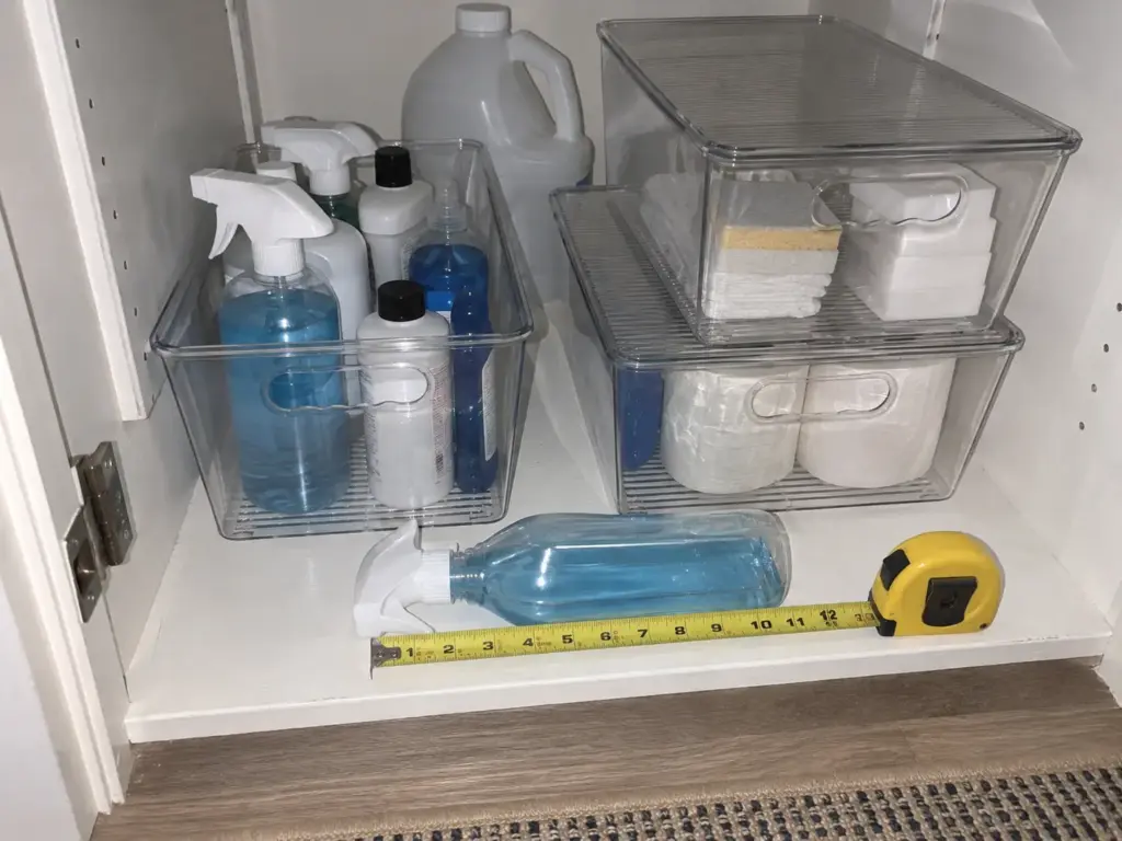 6 Cleaning Supply Organization Ideas That Stop Rebuying 6 Clear stackable bins with lids hold refill bottles and a bulk jug on the lowest closet shelf next to a tape measure